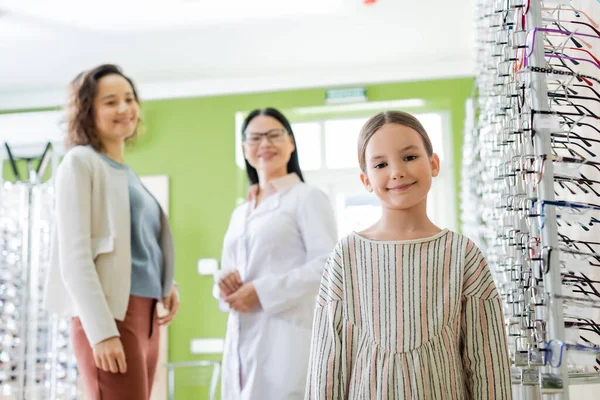 Cheerful girl looking at camera near blurred mom and asian ophthalmologist in optics store — Stockfoto