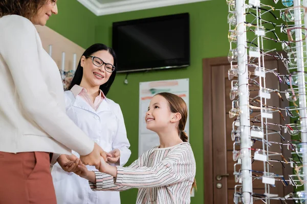 Happy girl holding hands with mom near happy asian oculist in optics shop — Stockfoto