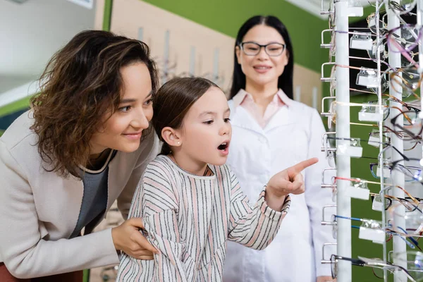 Amazed girl pointing at eyeglasses near smiling mom and blurred asian oculist in optics salon — Foto stock