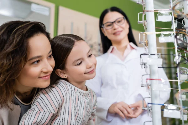 Smiling girl choosing eyeglasses near happy mom and blurred asian oculist in optics salon — Stock Photo