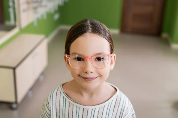 Fille heureuse dans les lunettes de vue regardant la caméra dans le magasin d'optique sur fond flou — Photo de stock