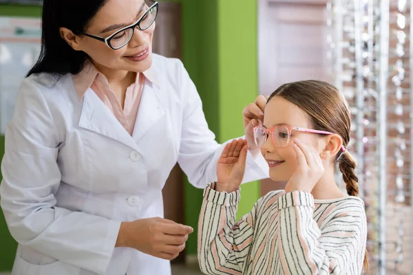 Smiling asian oculist choosing eyeglasses for girl in optics shop — Stock Photo