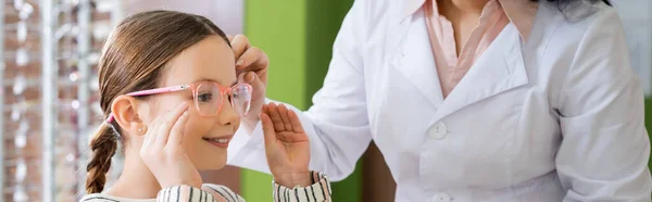 Positive girl choosing eyeglasses near ophthalmologist in optics salon, banner — Fotografia de Stock