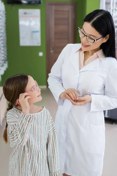 Asian oculist smiling at girl trying on eyeglasses in optics shop — Stock Photo