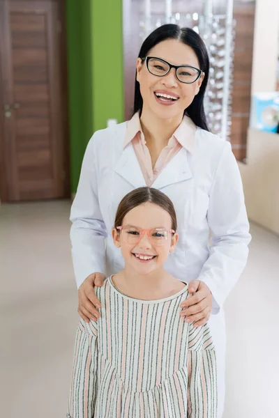 Cheerful asian oculist and girl in eyeglasses looking at camera in optics shop - foto de stock