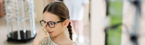 Chica positiva ajustando las gafas en la tienda de óptica sobre fondo borroso, bandera - foto de stock