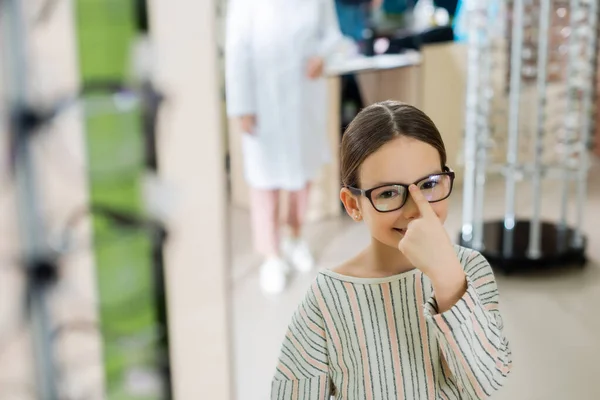 Cheerful girl looking at camera while adjusting eyeglasses in optics shop on blurred foreground — стоковое фото