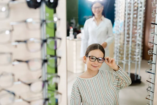 Pleased girl adjusting eyeglasses near blurred asian ophthalmologist in optics store — Foto stock