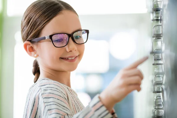 Усміхнена дівчина в окулярах вказує пальцем в магазині оптики на розмитому передньому плані — стокове фото