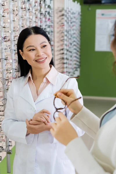 Blurred woman choosing eyeglasses near smiling asian ophthalmologist in optics shop — Foto stock