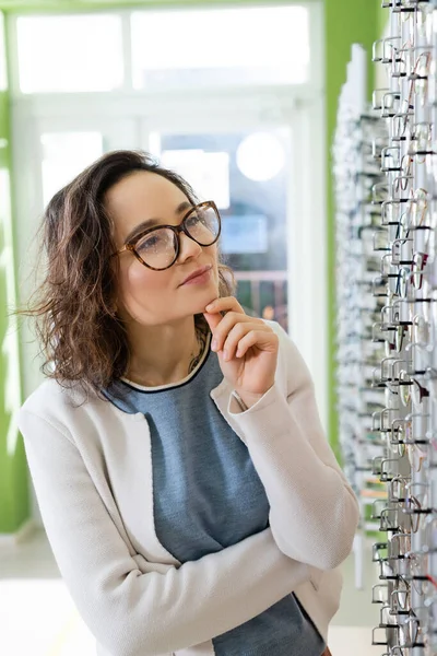 Thoughtful woman choosing eyeglasses in optics store on blurred background — стоковое фото