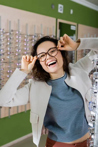 Excité femme rire à la caméra tout en essayant sur les lunettes dans magasin d'optique — Photo de stock