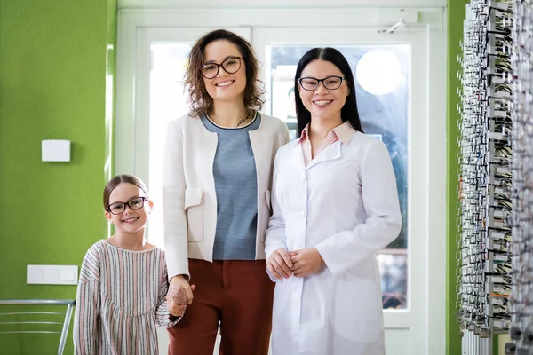 Happy asian oculist and mother with daughter smiling at camera in optics shop — Fotografia de Stock