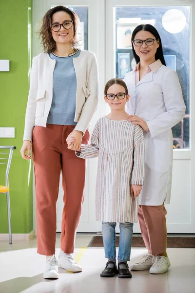 Mother and daughter with asian ophthalmologist in eyeglasses smiling at camera in optics shop — стоковое фото