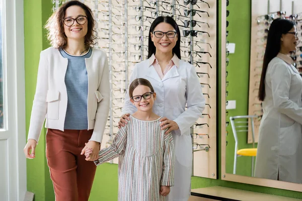 Happy mother, daughter and asian oculist in eyeglasses looking at camera in optics salon — стоковое фото