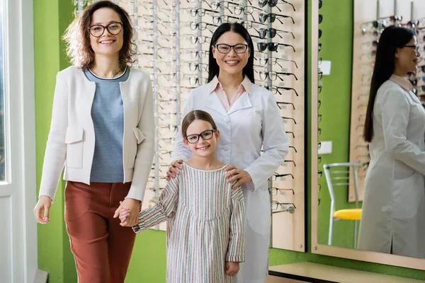 Happy mother, daughter and asian oculist in eyeglasses looking at camera in optics salon — Stock Photo
