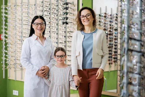 Happy girl in eyeglasses holding hands with mom and asian oculist while looking at camera in optics salon — Stock Photo