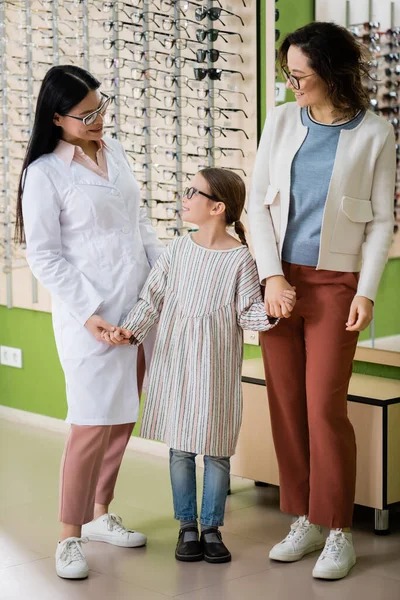 Feliz chica en gafas cogidas de la mano con madre y asiático oculista en óptica tienda - foto de stock