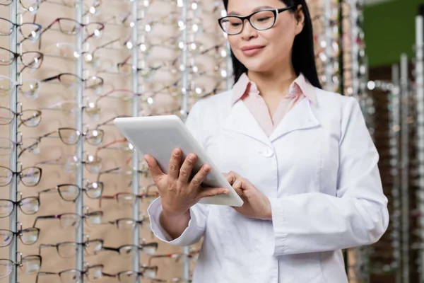 Asian ophthalmologist in eyeglasses using digital tablet while working in optics store — стоковое фото