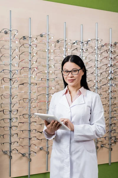 Happy asian oculist looking at camera while using digital tablet in optics salon — Stockfoto