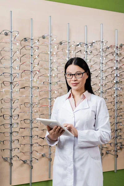 Happy asian oculist looking at camera while using digital tablet in optics salon — Stock Photo