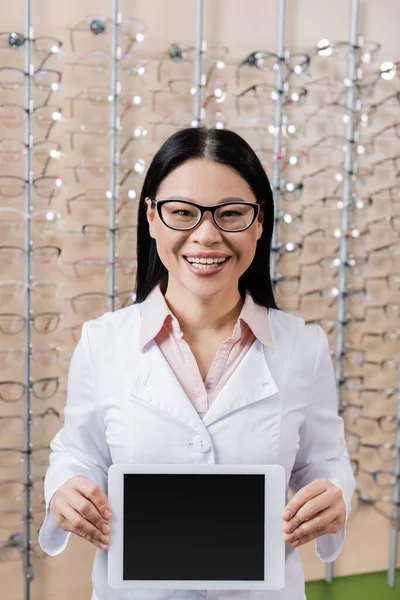 Happy asian oculist in eyeglasses holding digital tablet with blank screen in optics shop — Stockfoto