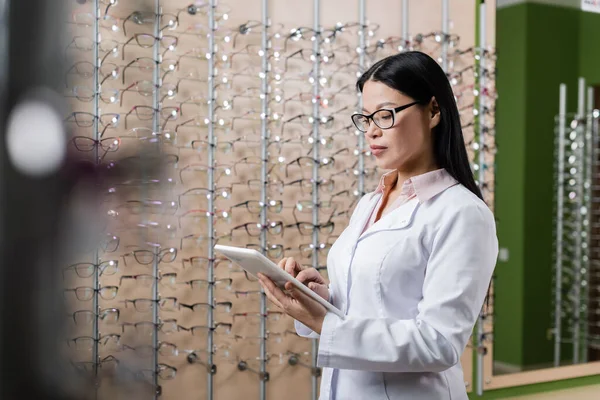 Asian ophthalmologist in eyeglasses using digital tablet in optics shop on blurred foreground — Stock Photo