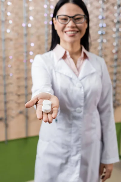 Alegre asiático oculista en gafas celebración lente caso en extendido mano en óptica tienda - foto de stock