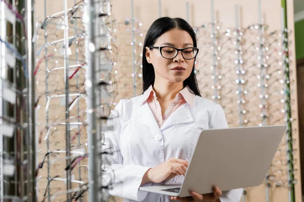 Asian ophthalmologist using laptop near assortment of eyeglasses in optics shop — Stock Photo