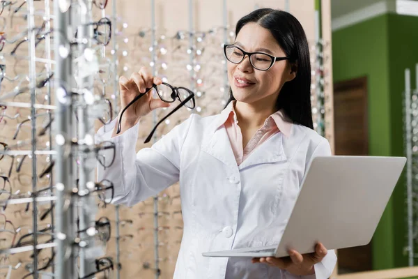 Pleased asian oculist with laptop looking at eyeglasses in optics salon — Stock Photo