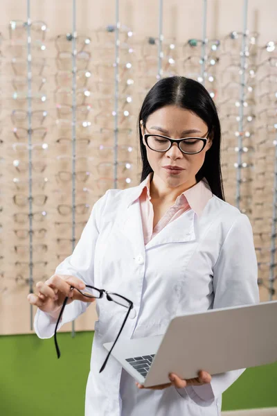 Asian opthalmologist holding laptop and eyeglasses while working in optics store — Stock Photo