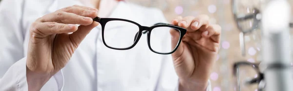 Vista recortada de oculista en abrigo blanco sosteniendo gafas en el salón de óptica, pancarta - foto de stock