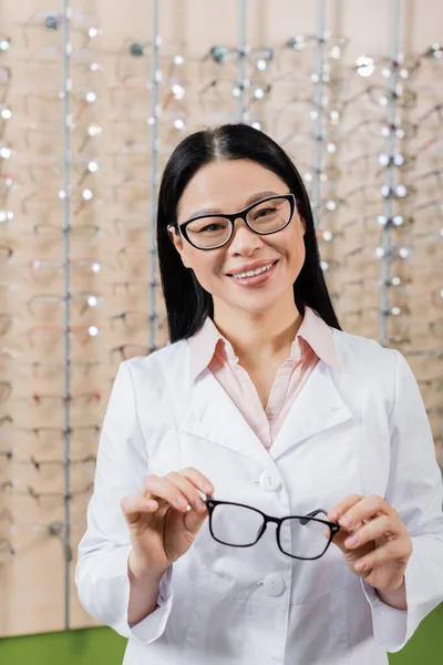 Cheerful asian ophthalmologist looking at camera while holding eyeglasses in optics store — Stock Photo
