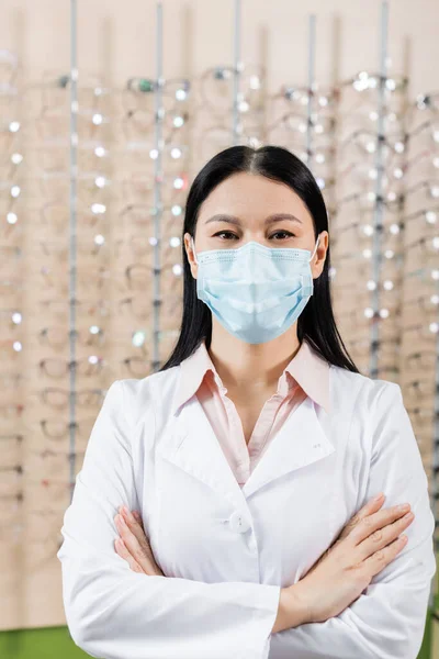 Asian ophthalmologist in medical mask standing with crossed arms in optics salon — Stock Photo