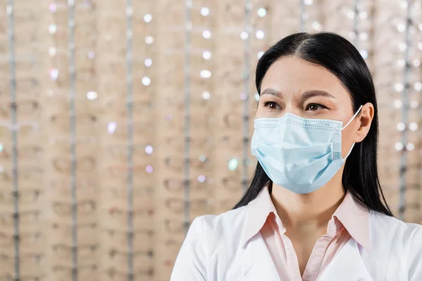 Asian oculist in medical mask near blurred assortment of eyeglasses in optics salon — Fotografia de Stock