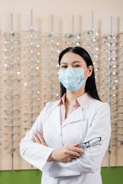 Asian ophthalmologist in medical mask standing with crossed arms near blurred assortment of eyeglasses in optics store — Stock Photo