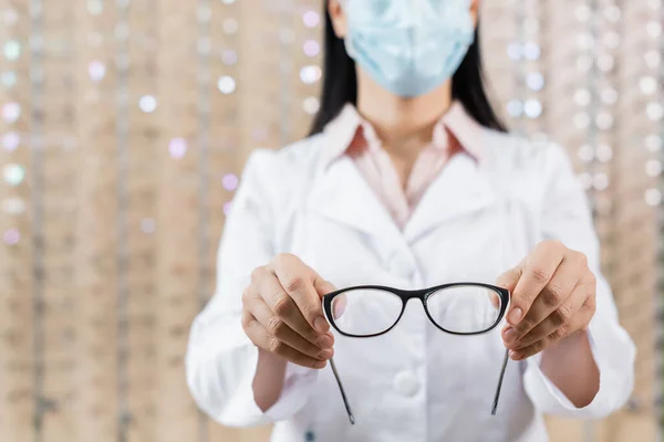 Cropped view of ophthalmologist in medical mask showing eyeglasses in optics store - foto de stock