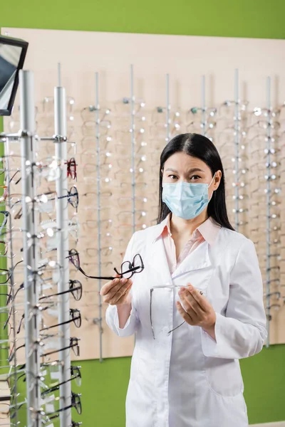 Asiático oculista en médico máscara comparar gafas mientras trabajo en óptica tienda - foto de stock