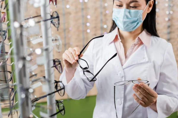 Asian ophthalmologist in medical mask comparing eyeglasses in optics shop — Stock Photo