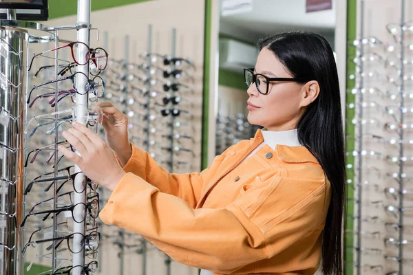 Asian woman choosing eyeglasses in optics salon on blurred background — Stockfoto