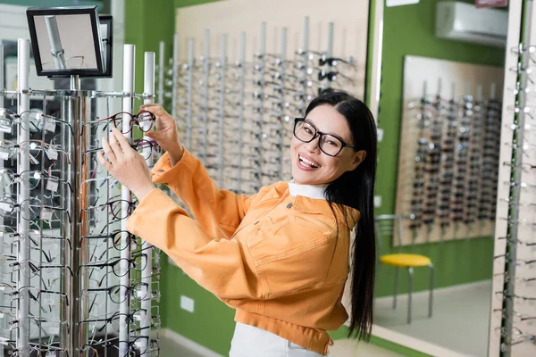 Joyful asian woman smiling at camera while choosing spectacles in optics store — Stock Photo