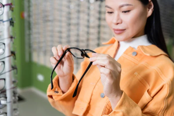 Asian woman choosing eyeglasses in optics store on blurred background — стоковое фото