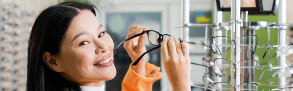 Happy asian woman looking at camera while choosing eyeglasses in optics salon, banner — Stock Photo