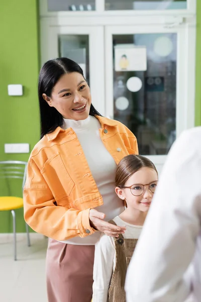 Cheerful asian woman talking to blurred ophthalmologist near daughter in eyeglasses in optics shop — Stock Photo