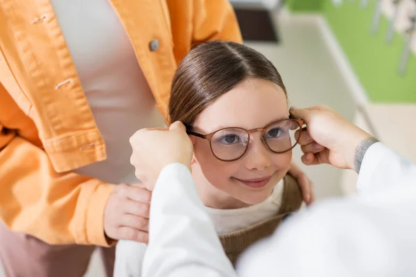 Oftalmólogo borrosa poner gafas en chica positiva en la tienda de óptica - foto de stock