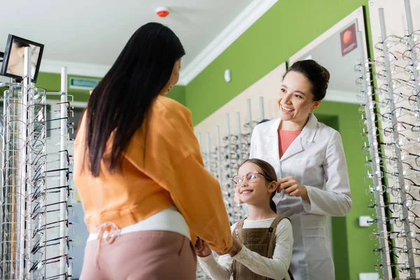 Happy girl in eyeglasses holding hands with mom near smiling oculist in optics shop — Stock Photo