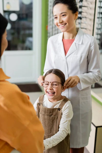 Positive oculist trying eyeglasses on cheerful girl near blurred mother in optics store — стоковое фото