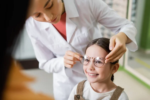 Ophthalmologist trying eyeglasses on smiling girl near blurred mom in optics store — Stock Photo