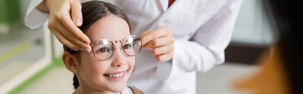 Oculista intentar gafas en sonriente chica cerca borrosa madre en óptica tienda, pancarta - foto de stock