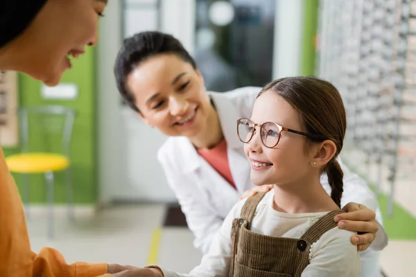 Feliz chica en gafas sonriendo cerca de asiático madre y borrosa oculista en óptica tienda - foto de stock